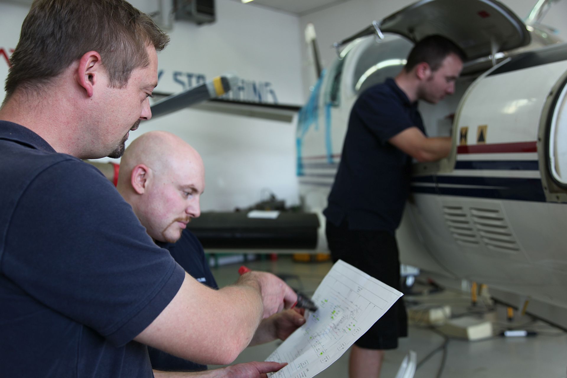 Techniker beim Einbau eines Gerätes in ein Flugzeug. Im Vordergrund zwei Männer, die einen Schaltplan betrachten, im Hintergrund ein Techniker, der einen geöffneten Klappe eines Flugzeuges steht.