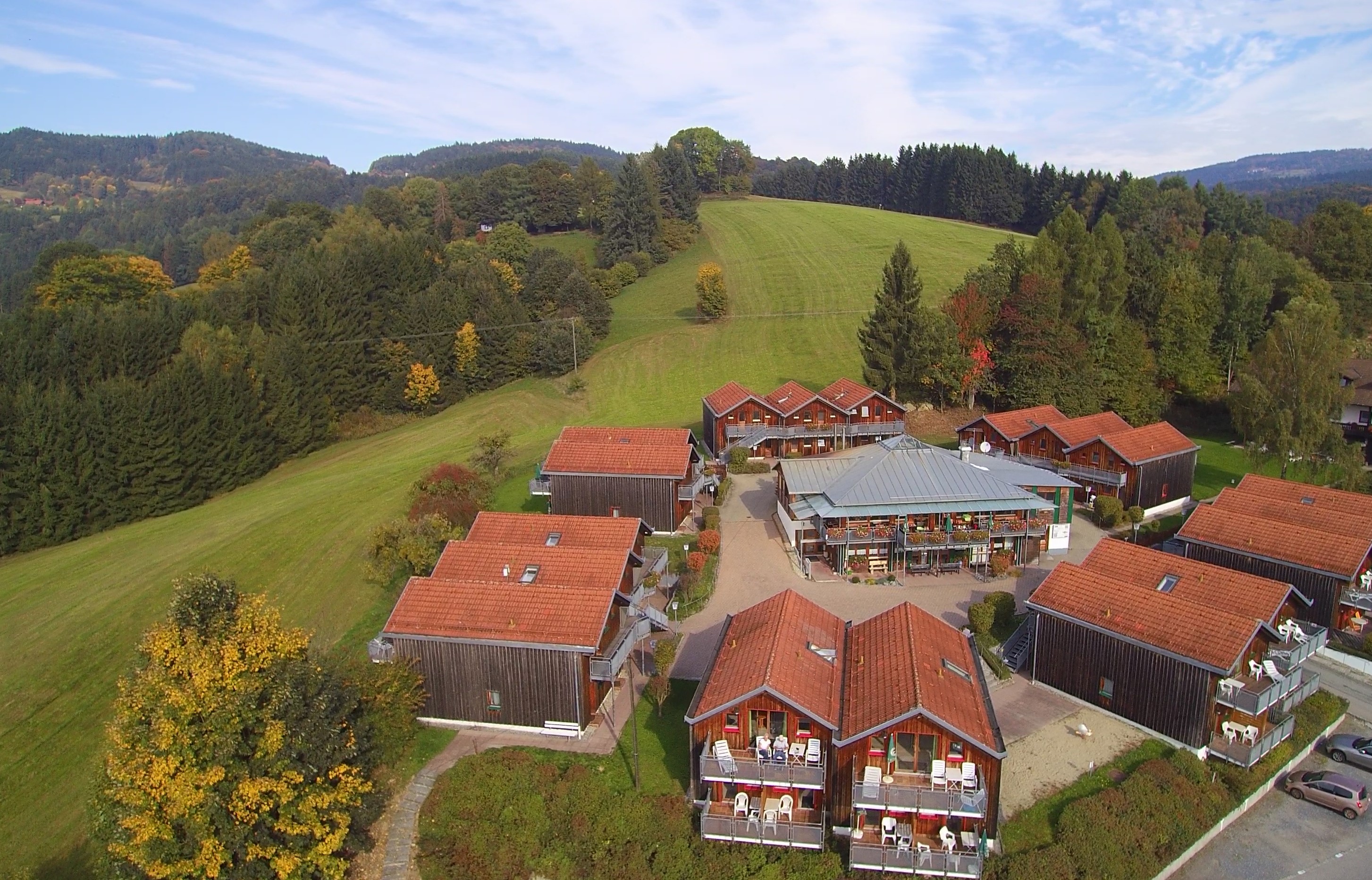 Luftbildaufnahme des Village Hotels Bayerischer Wald. Um das Hauptgebäude in der Mitte gruppieren sich mehrere Gebäude mit Holzverkleidung. In diesen befinden sich die Zimmer.